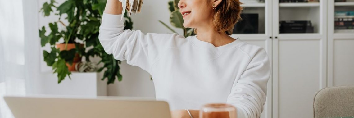Smiling woman holding cash, working at home office with a laptop, symbolizing financial success.