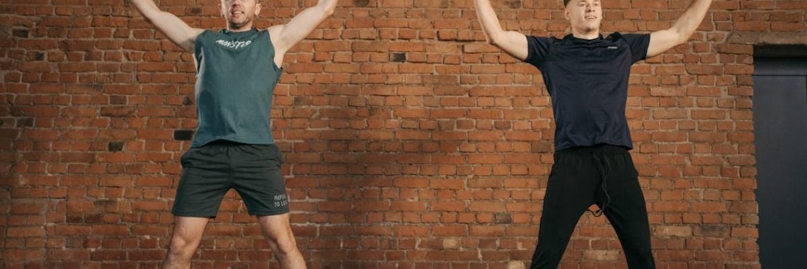 Two men performing jumping jacks in a gym setting, showcasing fitness and energy.