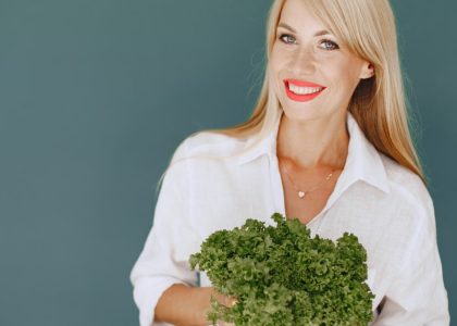 Cheerful woman with blonde hair holding fresh lettuce, promoting healthy living in a studio setting.